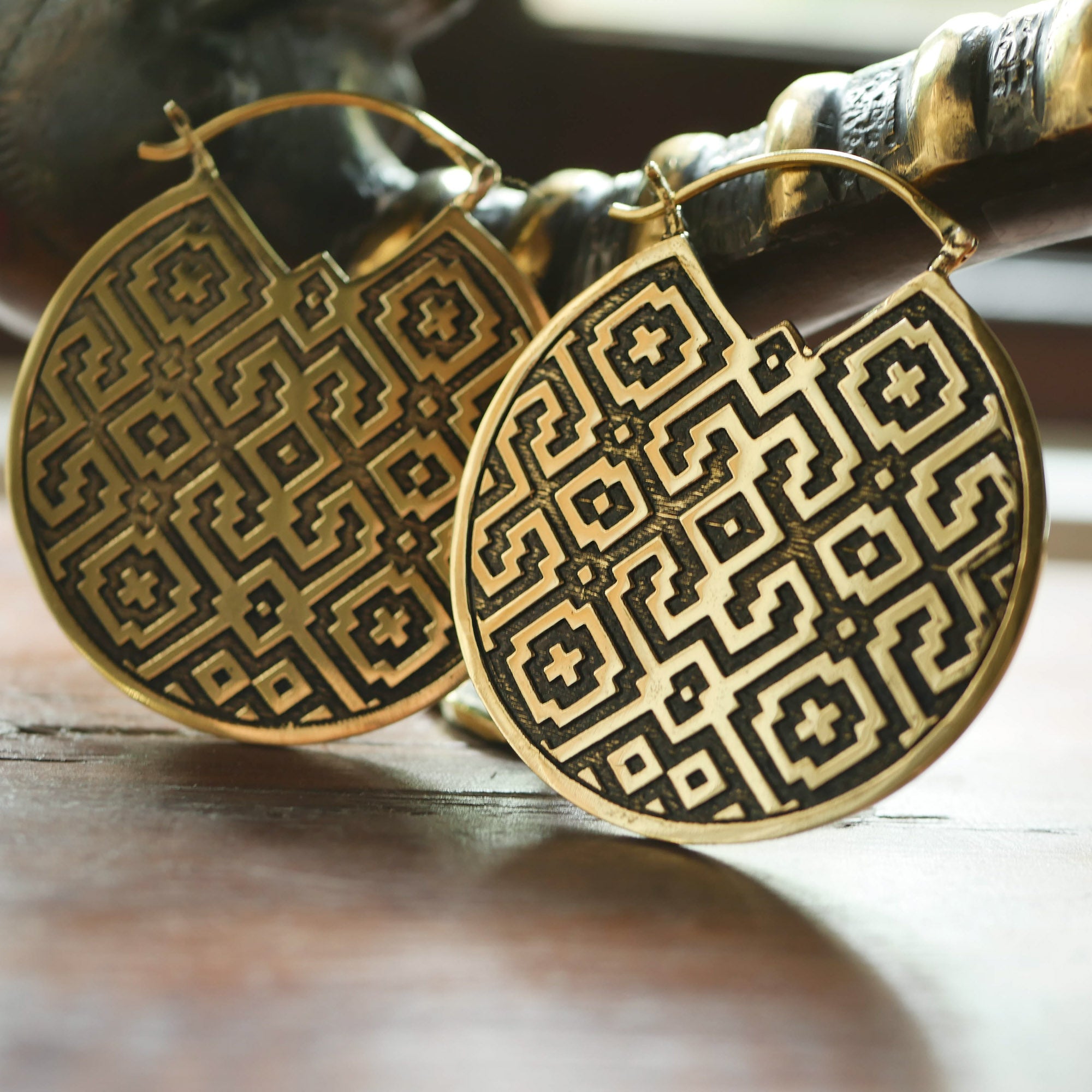 Close-up of the AREVALO Shipibo Oversized Disc Hoop Earrings in Gold by Kaya Movement, featuring intricate Shipibo patterns in black. The 14-gauge tribal disc hoop earrings are leaning against a wooden surface, with soft, natural light illuminating the details.